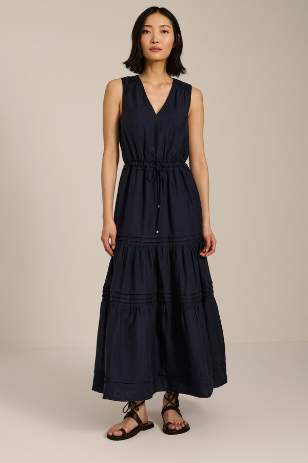 Woman wearing sleeveless navy maxi dress with V-neckline, drawcord waist, and pleated tiered skirt.