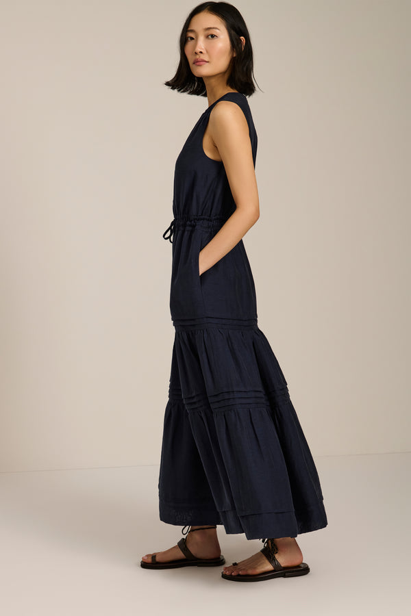 Woman wearing sleeveless navy maxi dress with V-neckline, drawcord waist, and pleated tiered skirt.