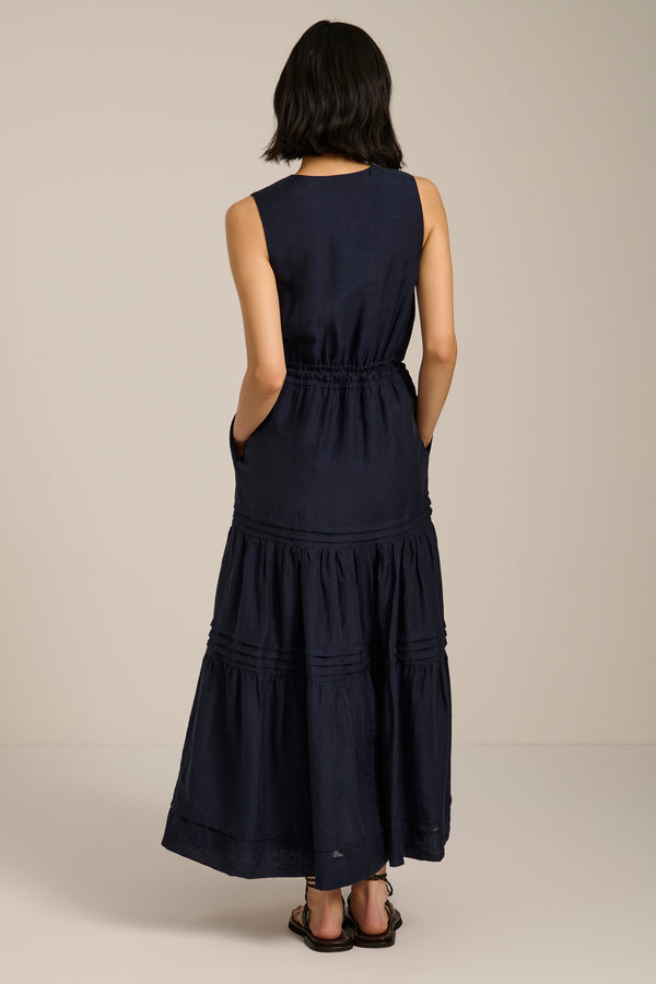 Woman wearing sleeveless navy maxi dress with V-neckline, drawcord waist, and pleated tiered skirt.