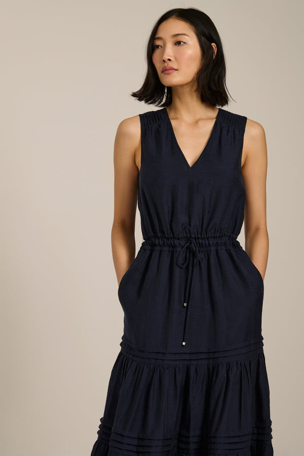 Woman wearing sleeveless navy maxi dress with V-neckline, drawcord waist, and pleated tiered skirt.